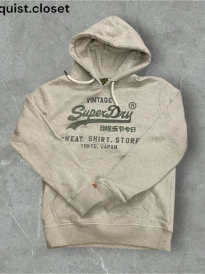 Grå hoodie från Superdry - Snygg beige hoodie från Superdry med tryck i grönt och svart på bröstet. Klassisk huva med vita dragsnören och känguruficka framtill. Tillverkad i mjukt bomullsmaterial, perfekt för en avslappnad streetwear-look. Det står storlek M men den är ganska liten i storlek så passar bättre som storlek S.