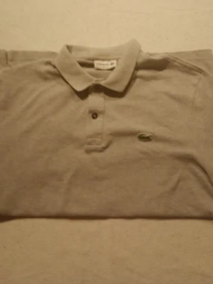 Beige pikétröja från Lacoste - Klassisk beige pikétröja från Lacoste med krage och två knappar framtill. Tröjan har den ikoniska gröna krokodilloggan broderad på bröstet. Perfekt för en stilren och avslappnad look.