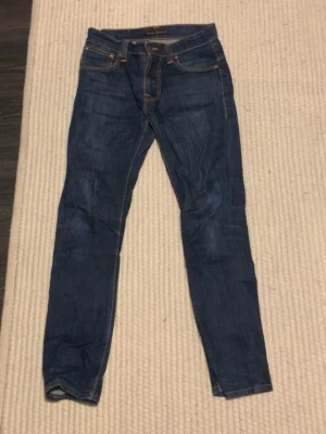 Mörkblå jeans från Nudie Jeans - Snygga mörkblå jeans från Nudie Jeans med klassisk femficksdesign och kontrastsömmar. Jeansen har raka ben och tydliga Nudie-sömmar på bakfickorna. Perfekta för en avslappnad och stilren look.