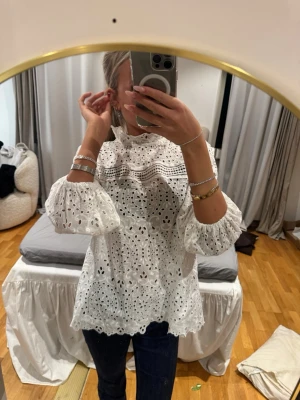 Vit broderad blus med volang - Superfin vit blus med broderade hålmönster och volangdetaljer vid hals och ärmslut. Blusen har en lös passform och är gjord i ett lätt bomullstyg som känns luftigt. Perfekt att styla med jeans eller kjol för en trendig look.