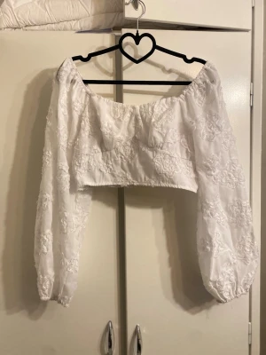 Vit croppad blus med broderade blommor - Supersöt vit croppad blus med puffiga långärmar och broderade blommor. Offshoulder-modell som ger en romantisk vibe och passar perfekt till höga jeans eller kjol. Tunn och luftig, perfekt för varmare dagar.