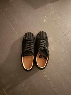 Svarta sneakers med nitar och röd sula - Säljer ett par svarta sneakers från Christian Louboutin med coola nitar över hela tån och klassisk röd sula. Skorna har snörning och är tillverkade i mocka för en lyxig känsla. Kontakta gärna innan köp💕.