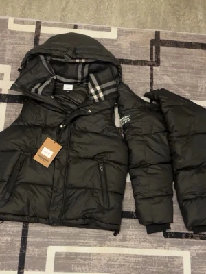 Svart dunjacka från Burberry - Snygg svart dunjacka från Burberry med avtagbar huva och klassiskt rutigt foder. Jackan har dragkedja framtill, två sidofickor med dragkedja och diskret Burberry-logga på ärmen. Perfekt för kalla dagar och riktigt trendig look.  Exklusiv Burberry-jacka av hög kvalitet som enkelt kan användas både som jacka och väst tack vare avtagbara ärmar. Designad med Burberrys klassiska rutmönster på insidan för en tidlös och elegant stil.  Storlek M 