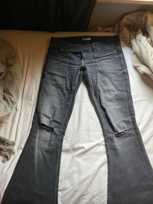Grå bootcut jeans från Gina tricot - Snygga grå jeans från Gina tricot med bootcut passform och slitningar vid knäna. Jeansen har dubbla knappar i midjan, framfickor och bälteshällor. Perfekta för en trendig och avslappnad stil.
