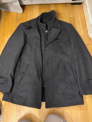 Mörkgrå ullrock med quiltad insida - Snygg mörkgrå rock i ullmix med klassisk krage och dold dragkedja. Jackan har tre fickor med knappdetaljer och quiltad insida för extra värme. Perfekt för kyliga dagar och stilrena outfits.