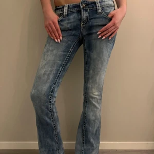 Blå Miss Me bootcut jeans - Snygga blå jeans med slitna detaljer och dekorativa bakfickor med broderi och knappar. Modellen har låg midja och bootcut passform. Jeansen är tillverkade i klassiskt denimtyg och har tydliga kontrastsömmar. 