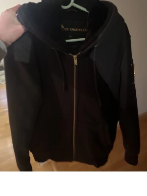 Mosse knuckels Bunny  - Snygg svart hoodie från Moose Knuckles med dragkedja framtill och guldfärgade detaljer. Utrustad med stor huva och quiltat innerfoder för extra komfort. Praktiska fickor och en cool patch på ärmen. Perfekt för dig som vill ha en stilren och exklusiv look.äkta 