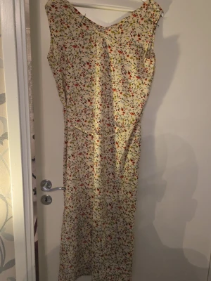 Blommig långklänning i ljusa toner - Söt långklänning med blommigt mönster i rött, grönt och gult på ljus botten. Ärmlös modell med v-ringning både fram och bak. Perfekt för varma dagar och har en avslappnad passform. Materialet känns lätt och luftigt, troligen bomull eller polyester.