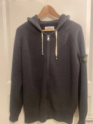 Grå stickad hoodie från Stone Island - Mörkgrå stickad hoodie från Stone Island med dragkedja framtill och vit snörning i huvan. Klassisk logga på vänster ärm. Tröjan har ribbade muddar och är långärmad, perfekt för kyliga dagar.