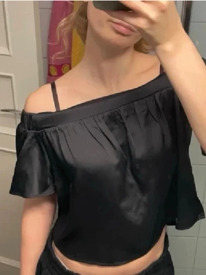 Svart offshoulder topp i satin - Säljer en svart offshoulder topp i glansigt satinliknande material. Toppen har korta, vida ärmar och elastisk kant upptill som gör att den sitter snyggt över axlarna. Den är croppad och har tunna axelband som går att knäppa av.