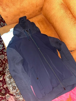 Mörkblå cardigan med huva från herno  - Snygg mörkblå cardigan med huva och hel dragkedja framtill. Jackan har ribbade muddar, snörning i huvan och två fickor med knappstängning. Perfekt för blåsiga dagar och enkel att matcha till olika outfits. Kvitto,tag finns, helt oanvänd 