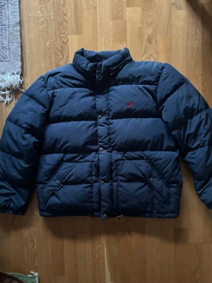 Mörkblå pufferjacka från Polo Ralph Lauren - Snygg mörkblå pufferjacka från Polo Ralph Lauren med klassisk röd logga på bröstet. Jackan har hög krage, tryckknappar och dragkedja samt två stora fickor framtill. Perfekt för kalla dagar och riktigt stilren look.