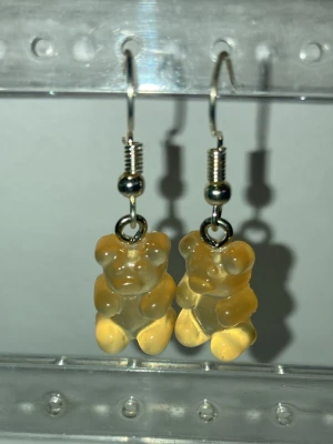 Guldbjörnsörhängen i akryl - Handgjorda örhängen av mig med genomskinliga guldfärgade björnar i akrylplast. Hängande modell med silvriga krokar och detaljer. Perfekt accessoar för dig som gillar lekfull och unik stil. Frakt 22-100kr. Kontakta mig om du har frågor eller vill köpa.