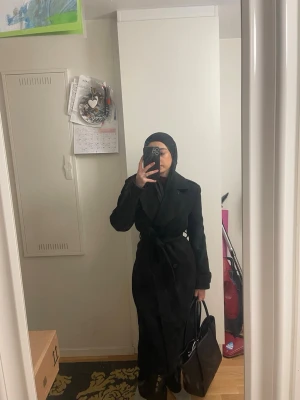 Svart lång trenchcoat med bälte - Stilren svart trenchcoat med klassisk krage och knytskärp i midjan. Lång modell med markerad midja och långa ärmar. Perfekt för en trendig och elegant look.