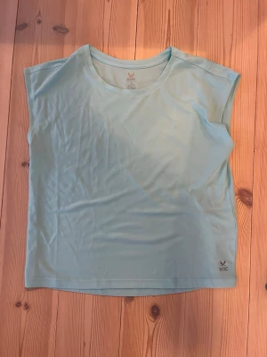 Ljusblå sporttopp från SOC - Säljer en ljusblå sporttopp från SOC i tekniskt funktionsmaterial. Toppen har rund halsringning, korta ärmar och en luftig passform som passar perfekt till träning. Diskret SOC-logga nere vid fållen. Enkel och stilren design.