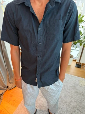 Mörkblå kortärmad skjorta - Stilren mörkblå skjorta med korta ärmar och klassisk krage. Skjortan har en bröstficka och knäppning framtill. Perfekt för en avslappnad men snygg look. Tillverkad i mjuk bomull som känns skön mot huden.