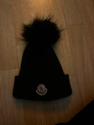 Svart Moncler mössa med pälsboll - Svart ribbstickad mössa från Moncler med stor fluffig pälsboll på toppen och klassisk Moncler-logga framtill. Mössan har uppvikt kant och är riktigt varm och snygg till vintern.