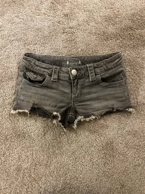 Grå low waist korta jeansshorts - SKRIV PRIVAT FÖR MER BILDER!! PRIS KAN DISKUTERAS! inte lika små sen fem ser ut på bilden!Storlek xs! Säljer ett par grå denimshorts med rå, fransig kant och klassiska fem fickor. Shortsen har bälteshällor, knappgylf och bakfickor med lock och knapp. Perfekta för sommardagar och en avslappnad look.