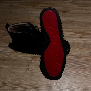 Svarta sneakers med nitar och röd sula - Säljer ett par svarta high top sneakers från Christian Louboutin i mocka med svarta nitar på sidorna och klassisk röd sula. Skorna har snörning och en chunky look som sticker ut. Perfekta för dig som vill ha något edgy och exklusivt.
