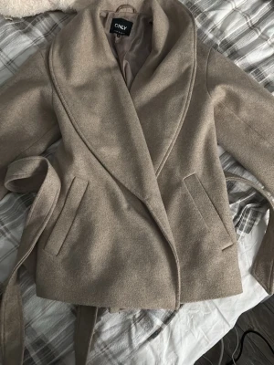 Beige omlottkappa från ONLY - Snygg beige kappa från ONLY med omlottmodell och brett slag. Kappan har bälte i midjan och två fickor framtill. Perfekt för dig som gillar en stilren och enkel look. Materialet känns mjukt och har en ullig känsla.