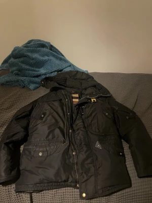 Svart vinterjacka från Parajumpers - Säljer en svart vinterjacka från Parajumpers med huva, flera fickor och gul detalj vid halsen. Jackan har dragkedja och knappar framtill samt en klassisk passform. Perfekt för kalla dagar och riktigt snygg streetstil.