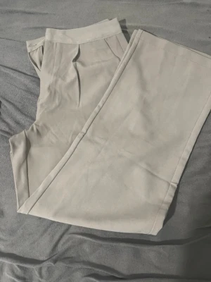 Beige kostymbyxor med pressveck - Snygga beige kostymbyxor med raka ben och klassiska pressveck framtill. Byxorna har hög midja och är tillverkade i ett mjukt material som ger en clean och stilren look. Perfekta att matcha med en skjorta eller stickad tröja.