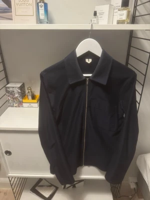 Mörkblå overshirt från arket  - Snygg mörkblå overshirt från arket med klassisk krage och dragkedja framtill. Jackan har en bröstficka och är långärmad. Perfekt för lager på lager och enkel att matcha med olika outfits.
