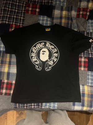 Svart Chrome Hearts x BAPE t-shirt XL - Säljer en svart t-shirt från Chrome Hearts x BAPE i storlek XL. Stor vit print fram med klassisk Chrome Hearts-logga och BAPE-apa, samt stor ryggprint med Baby Milo och kors. Skön passform och riktigt clean streetwear-vibe. Tröjan ser bra ut men använd några gånger. Inga defekter