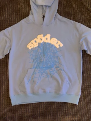 Ljusblå hoodie med sp5der-tryck - Säljer en ljusblå hoodie från Sp5der med stor ficka framtill och huva. På bröstet finns ett vitt sp5der-tryck och ett blått spindelnätsmotiv. Hoodien har ribbade muddar och är gjord i mjukt bomullsmaterial. Perfekt för dig som gillar streetwear och unika prints.