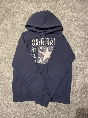 Mörkblå hoodie med stjärntryck - Snygg mörkblå hoodie med huva och tryck framtill som visar en stor stjärna och texten 'ORIGINAL FIT EST. 2024'. Hoodien har känguruficka och ribbade muddar vid ärmar och nederkant. Perfekt för en avslappnad och cool stil.