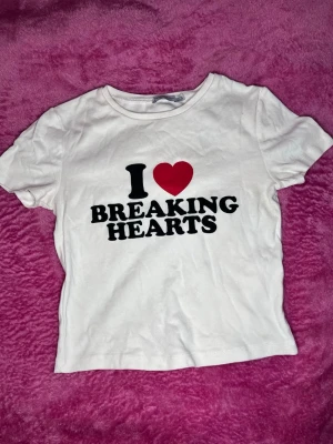 Vit t-shirt med tryck och hjärta - Vit t-shirt med svart text och ett rött hjärta på bröstet där det står 'I ♥ BREAKING HEARTS'. Klassisk rund halsringning och korta ärmar. Perfekt statement-plagg för dig som gillar att sticka ut.