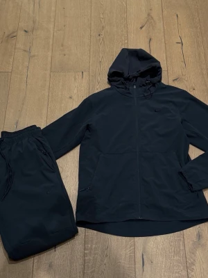 Svart Nike träningsset med huva - Snyggt svart träningsset från Nike med matchande jacka och byxor. Jackan har huva, dragkedja och två fickor framtill. Byxorna har resår i midjan och dragsko för bästa passform. Båda plaggen har diskret Nike-logga. Perfekt för träning eller chill.