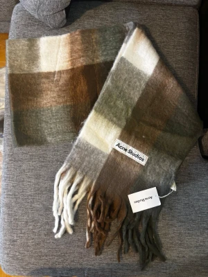 Rutig halsduk från Acne Studios - Säljer en supermjuk och fluffig halsduk från Acne Studios i rutigt mönster med nyanser av brunt, beige, grått och grönt. Halsduken har långa fransar i ändarna och en tydlig Acne Studios-logga. Perfekt accessoar för att lyfta din outfit. Mått 220x40 cm