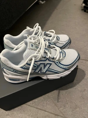 Vita New Balance sneakers med blå detaljer - Snygga vita New Balance sneakers med blå detaljer och mesh-överdel för extra andningsförmåga. Klassisk N-logga på sidan, snörning och bekväm platt sula. Perfekta för dig som gillar sportig och stilren look.