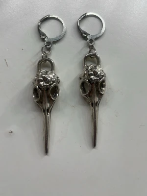 Handgjorda örhängen med fågelskalle - Unika silvriga örhängen i form av fågelskallar med detaljerade utskärningar. Hängande modell med ringfäste, tillverkade i metall. Perfekta för dig som gillar alternativ stil och vill sticka ut med accessoarerna.