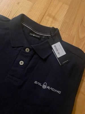 Marinblå pikétröja från Sail Racing - Snygg marinblå pikétröja från Sail Racing med klassisk krage och två knappar framtill. Tröjan har broderad logga på bröstet och är gjord i ett mjukt, pikévävt material. Perfekt för en stilren och sportig look.