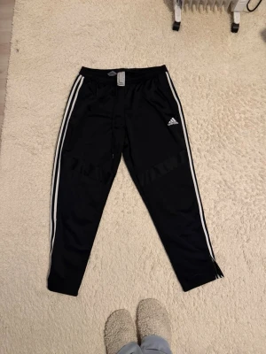 Svarta Adidas mjukisbyxor med vita ränder - Snygga svarta mjukisbyxor från Adidas med klassiska vita ränder längs sidorna och Adidas-logga på låret. Byxorna har snörning i midjan och är tillverkade i polyester för en sportig och bekväm känsla. Perfekta för chill eller träning. Säljer för 100kr plus frakt 