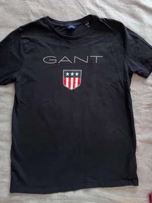 Svart GANT t-shirt med tryck - Snygg svart t-shirt från GANT med stort tryck framtill. T-shirten har korta ärmar och rund halsringning. På bröstet finns GANT-loggan och ett sköldmärke med stjärnor och ränder i vitt och rött. Perfekt till jeans eller shorts.