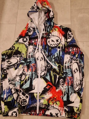 Färgstark graffiti hoodie med dragkedja - Säljer en hoodie med grymt färgstarkt graffiti-inspirerat tryck i blått, rött, grönt, svart och vitt. Hoodien har hel dragkedja, huva med snörning och coola tecknade motiv som sprayburkar och spelkontroller. Polyester. Perfekt för dig som vill sticka ut i mängden.