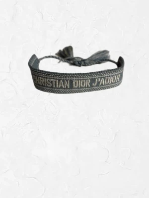 Christian Dior J'Adior armband - Snyggt grått tygarmband från Christian Dior med texten 'J'ADIOR' broderad i vit. Armbandet har justerbar knytning med tofsar i ändarna.