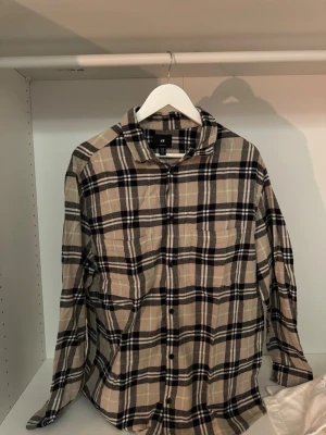 Beige och svart rutig skjorta H&M - Klassisk rutig skjorta från H&M i beige och svart med regular fit. Skjortan har långa ärmar, knappar framtill och mjukt bomullsmaterial. Perfekt för en avslappnad och trendig look.