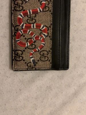 Gucci korthållare med orm och GG-mönster - Snygg korthållare från Gucci i beige och svart med klassiskt GG-mönster och en röd, vit och svart orm på framsidan. Tillverkad i skinn med plats för flera kort. Perfekt accessoar för dig som vill ha något unikt och trendigt.