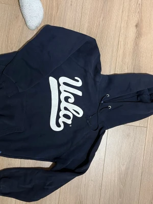 Marinblå UCLA hoodie med tryck - Snygg marinblå hoodie från UCLA med stort vitt tryck på bröstet och ett emblem på huvan. Klassisk känguruficka framtill och dragsko i huvan. Perfekt för en avslappnad och sportig stil.