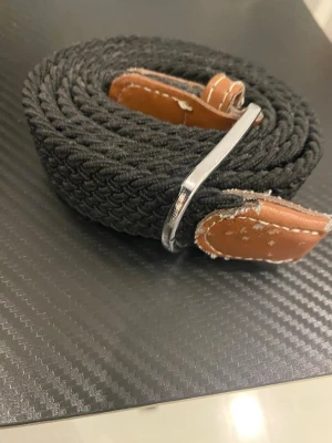 Svart flätat bälte med läderdetaljer - Snyggt svart flätat bälte med bruna läderdetaljer och silverfärgat spänne. Bältet har en flexibel och stretchig design som gör det enkelt att justera. Passar perfekt till jeans eller chinos för en avslappnad men stilren look.