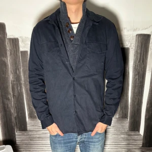 Mörkblå skjorta med knappar - Säljer en stilren mörkblå skjorta med knappar framtill och två bröstfickor. Skjortan har klassisk krage och lång ärm, perfekt att bära öppen över t-shirt eller stängd för en mer uppklädd look. Passar till jeans eller chinos.