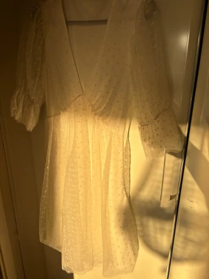 Vit kort prickig klänning med puffärm - Söt vit kort klänning med v-ringning och puffärmar. Klänningen har ett transparent lager med små prickar och voluminösa ärmar som ger en romantisk känsla. Perfekt för sommarens alla tillfällen och studentfirande.