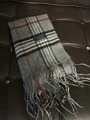 Grå rutig halsduk från Burberrys - Klassisk grå halsduk från Burberrys i 100% lammull med ikoniskt rutmönster i svart, vitt och röda detaljer. Halsduken har fransar i ändarna och en snygg vävd etikett. Perfekt accessoar för att lyfta din outfit.