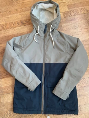 Tretorn vindjacka med teddyfoder - Snygg vinterjacka, camper jacket från Tretorn i tvåfärgad design, beige/grön upptill och svart nedtill. Jackan har huva med dragsko, hel dragkedja och mysigt teddyfoder på insidan. Perfekt för kyliga dagar och har praktiska fickor framtill. Jackan är sparsamt använd i mycket fint skick. Endast lite nopprig på insidan! 