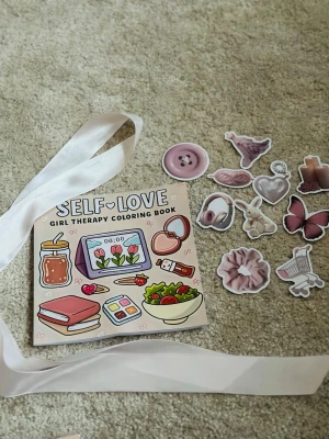 Self-Love Girl Therapy Coloring Book with stickers - En söt och inspirerande målarbok med temat self-love, fylld med illustrationer av vardagliga saker som böcker, sallad, hörlurar och smink. Omslaget är pastellfärgat med rosa och beige toner. Perfekt för dig som gillar att färglägga och pyssla.