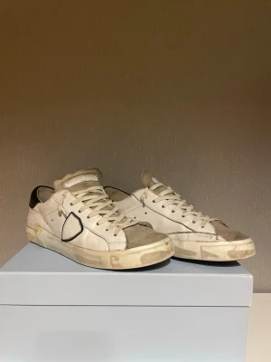 Philippe Model - Säljer ett par vita sneakers från Philippe Model med beige detaljer och svart häl. Skorna har snörning, rund tå och en platt sula. Materialmix av skinn och mocka ger en schysst kontrast. Klassisk Philippe Model-logga på sidan. Som ni ser på sista bilden så ska just denna modell se ut och vara lite sliten ut. Pris kan alltid diskuteras! 